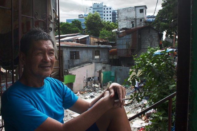 An old man on Kalayaan Avenue smokes a cigarette, with buildings from Bonifacio Global City in the background.